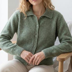 May include: A light green cardigan with a collared neckline and button-down front. The long-sleeved sweater is made of a textured knit fabric. The model is wearing a gold necklace and light-colored pants.