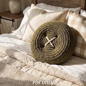 Puede incluir: Un cojín redondo de ganchillo color verde oliva con forma de botón grande y un cordón grueso de color crema cosido en forma de X en el centro. El texto PDF ONLY BEGINNER FRIENDLY está impreso en blanco en la parte inferior.
