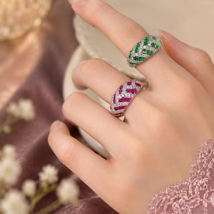 May include: Two silver-toned rings featuring a chevron pattern. One ring is set with vibrant green stones and clear white stones, while the other ring is set with bright pink stones and clear white stones, both arranged in a repeating zigzag design.