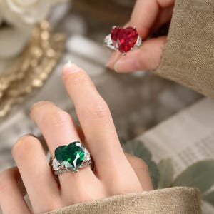 May include: Two rings featuring large heart-shaped gemstones, one in deep green and one in vibrant red. Each ring is set in a silver-toned metal band accented with smaller clear stones along the sides, showcasing a faceted cut and polished finish.