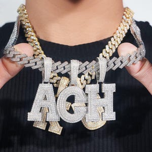 May include: Close-up of three diamond-encrusted pendant necklaces spelling out the letters "ACH". The pendants hang from thick, silver and gold Cuban link chains. The necklaces are displayed against a black background.