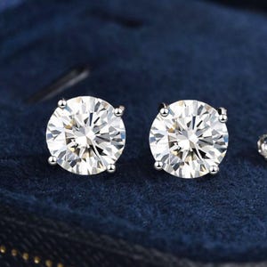 May include: A pair of round, clear gemstone stud earrings. The earrings are set in a silver-colored metal and are displayed against a dark blue background. The gemstones have a brilliant cut, reflecting light.