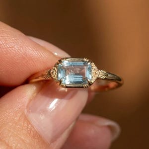 May include: A gold ring featuring a rectangular, light blue, emerald-cut gemstone set in a bezel setting. The metal band displays intricate, engraved leaf-like patterns on both sides of the central stone, creating a detailed, textured appearance.