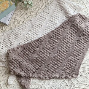 May include: Two triangular knitted shawls, one in cream and one in a muted mauve, featuring a repeating diagonal lace pattern with alternating sections of garter stitch and openwork eyelet rows, showcasing a soft texture and finished edges.
