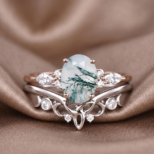 May include: A two-piece ring set featuring a silver band with a decorative design and a rose gold band with small clear stones. The main ring has an oval-shaped, light green stone with dark green veining.