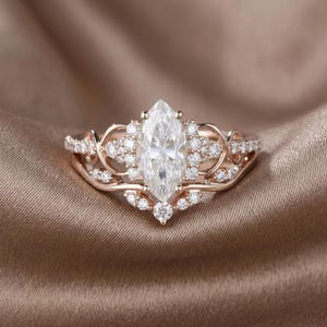 Puede incluir: Anillo de compromiso de oro rosa con una piedra central en forma de marquesa y diamantes redondos más pequeños. El anillo tiene un diseño intrincado con delicados filigranas y un halo de piedras más pequeñas alrededor del diamante principal.