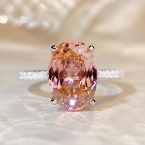 May include: A close-up of a ring featuring a large, oval-cut peach-colored gemstone. The gemstone is set in a silver band with smaller, clear stones. The ring is displayed on a soft, neutral background.