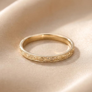 Può includere: Un anello a fascia d'oro con un intricato motivo floreale inciso all'esterno. L'anello è posto su uno sfondo morbido e neutro, che mette in risalto il suo design delicato e il colore caldo.
