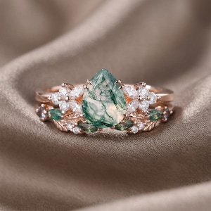 May include: A rose gold ring set featuring a pear-shaped moss agate gemstone. The ring is accented with white and green gemstones, and floral details. The set includes a matching band with similar floral and gemstone accents.