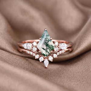 May include: A rose gold ring set featuring a central, kite-shaped moss agate gemstone with green inclusions. The engagement ring is paired with a delicate band adorned with small, round, and marquise-cut clear gemstones. The set is displayed on a neutral, textured surface.