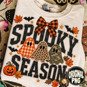 May include: A cream-colored crewneck sweatshirt featuring the text SPOOKY SEASON in black block letters. The design includes three ghosts with orange gingham, leopard print, and camouflage patterns, an orange plaid bow, bats, stars, leaves, and a pumpkin.