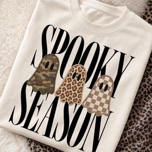 May include: A cream-colored crewneck sweatshirt featuring the black text SPOOKY SEASON. The design includes three ghost-shaped patches: one with a green camouflage pattern, one with a brown leopard print, and one with a tan and white checkerboard pattern.