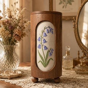 May include: A cylindrical wooden jewellery box standing on three small spherical feet. The front panel features an oval inset of cream-coloured fabric embroidered with a botanical design of blue bell-shaped flowers and green leaves on a stem.