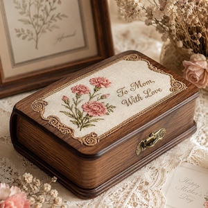 May include: A rectangular wooden box with a dark brown wood grain finish and a brass latch. The lid features an inset fabric panel embroidered with pink carnations and the text To Mom, With Love, framed by a decorative gold-toned border.