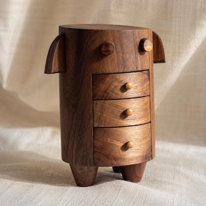 May include: A dark brown wooden chest of drawers featuring a cylindrical body, three stacked drawers with small round knobs, two protruding side panels, two circular top accents, and three short, sturdy legs supporting the base structure.