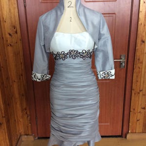 May include: A gray cocktail dress with a matching bolero jacket displayed on a mannequin. The dress features a ruched skirt and a white bodice with floral embroidery. The jacket has three-quarter sleeves with floral detailing.