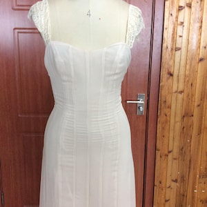 Può includere: Abito da sposa color crema su manichino. L'abito presenta un corpetto aderente con cuciture verticali e una gonna fluida. Delicati pizzi rifiniscono le maniche corte. L'abito è esposto davanti a una porta di legno.