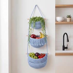 May include: A three-tiered hanging storage organizer made of light blue crocheted yarn and wooden dowels. The top basket holds a trailing green plant, the middle basket contains apples and bananas, and the bottom basket holds grapes and tomatoes.