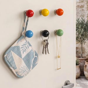 Könnte beinhalten: Ein Set aus sechs Wandhaken mit kugelförmigen Enden in Rot, Gelb, Orange, Blau, Schwarz und Grün. Ein Stoff-Topflappen mit blau-weißem Blattmuster hängt am blauen Haken, während Schlüssel und eine goldene Halskette an anderen Haken hängen.