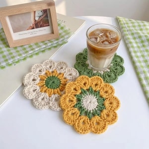 May include: Three round, flower-shaped crochet coasters featuring intricate petal designs. One coaster has a green center with yellow and cream petals, another is solid green, and the third has a cream center with green and yellow petals.