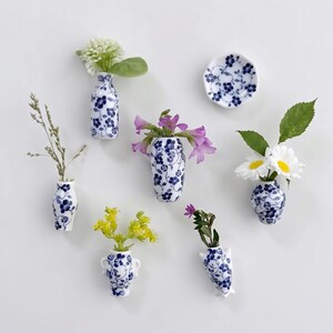 May include: A collection of six small ceramic wall-mounted vases and one small dish, all featuring a white base with a blue floral pattern. Each piece holds various small flowers, including white daisies, purple blossoms, and green foliage.