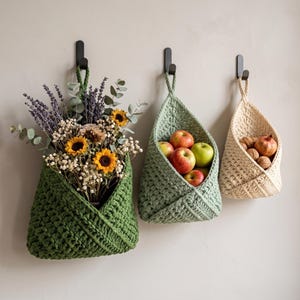 May include: Three hanging crochet baskets in dark green, sage green, and cream, each suspended from a black wall hook. The dark green basket holds dried flowers, the sage green basket contains red and green apples, and the cream basket holds nuts.