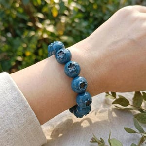 May include: A bracelet composed of round, deep blue beads that resemble blueberries, each featuring a small, star-shaped indentation on the surface. The beads are strung together in a continuous line, creating a textured, spherical appearance.
