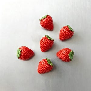 May include: Six small, three-dimensional strawberry-shaped charms or beads. Each piece features a bright red, textured surface with a pitted appearance and a small, molded green leafy stem at the top. The items are arranged in a scattered pattern.