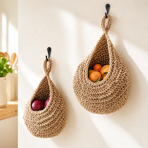 May include: Two teardrop-shaped hanging baskets made of woven tan jute fiber. The smaller basket on the left contains two red onions, while the larger basket on the right holds several orange citrus fruits. Both baskets hang from black metal hooks.