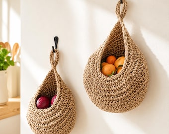 May include: Two teardrop-shaped hanging baskets made of woven tan jute fiber. The smaller basket on the left contains two red onions, while the larger basket on the right holds several orange citrus fruits. Both baskets hang from black metal hooks.