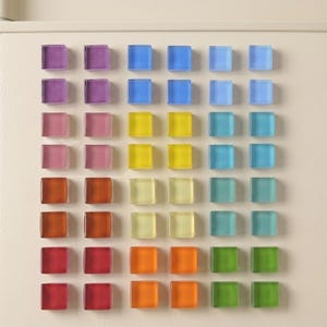 May include: A collection of square glass magnets arranged in a grid on a flat surface. The magnets feature a variety of translucent colors including purple, blue, yellow, pink, orange, green, and teal, organized in columns by color hue.