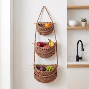 Peut inclure: Un organisateur de rangement suspendu à trois niveaux composé de paniers en crochet marron suspendus par des tiges en bois clair et une corde assortie. Les paniers contiennent une orange, des pommes rouges, des bananes, une prune et du raisin.