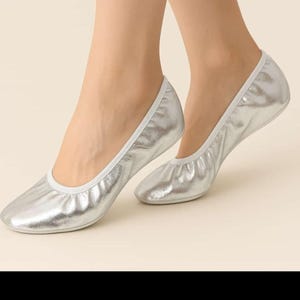 Könnte beinhalten: Silberne Ballerinas mit metallischem Glanz. Die Schuhe haben eine runde Spitze und eine weiße Einfassung. Die Ballerinas sind faltbar und werden an Füßen gezeigt.