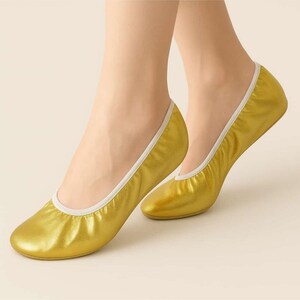 Könnte beinhalten: Ein Paar goldfarbene Ballerinas mit weißem Rand. Diese faltbaren Schuhe haben eine glänzende, metallische Oberfläche und eine runde Spitze. Die Ballerinas sind auf Komfort und Stil ausgelegt.