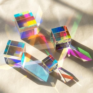 May include: Four clear glass cubes are arranged on a light surface, each refracting light into a spectrum of colors. The cubes vary in size, with the largest at the top left and the smallest at the bottom center, creating colorful shadows.