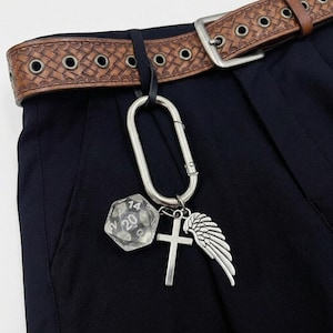 Puede incluir: Un cinturón de cuero marrón con textura tejida y hebilla plateada. Un mosquetón plateado sujeta un dado transparente de múltiples caras, una cruz plateada y un dije de ala plateada. El cinturón se usa con pantalones azul oscuro.