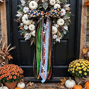 May include: A decorative wreath featuring white pumpkins, pinecones, and greenery, accented by a large bow with a candy corn pattern and long, multi-textured ribbons in green, orange, black, and white, hanging against a dark door.