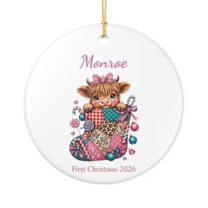 May include: A round white ceramic ornament featuring a fluffy Highland cow calf wearing a pink bow, peeking out of a colorful patchwork Christmas stocking. The text reads Monroe and First Christmas 2026, accented with candy canes and ornaments.