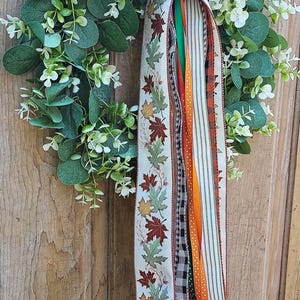 May include: A circular wreath featuring green eucalyptus leaves and small white flowers, accented by a large bow made of cream-colored ribbon with a multi-colored autumn leaf pattern. Long, trailing ribbons in orange, green, and plaid hang down.