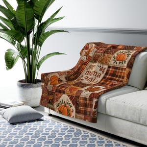 May include: A quilted throw blanket featuring a patchwork design of autumn leaves, plaid patterns, and orange pumpkins. The centre of the blanket displays the text "mama mini" in a brown script font, surrounded by fall-themed illustrations.