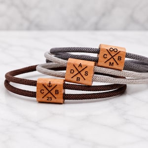 Könnte beinhalten: Drei Seilarmbänder mit quadratischen Holzakzenten. Die Armbänder sind in Braun-, Grau- und Hellgrautönen gehalten. Jeder Holzakzent hat ein anderes Design, einschließlich Initialen und Symbolen. Der Hintergrund ist eine weiße Marmoroberfläche.