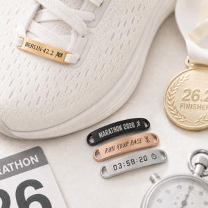 Könnte beinhalten: Ein weißer Sportschuh mit einem goldenen Schnürsenkel-Tag, graviert mit BERLIN 42.2 und einem Zielflaggen-Symbol. Drei weitere Metall-Tags in Schwarz, Roségold und Silber sind abgebildet, zusammen mit einer Goldmedaille und einer Stoppuhr.