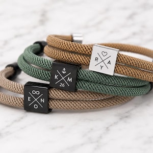 Könnte beinhalten: Drei mehrlagige Armbänder mit gewebten Kordelbändern in Beige und Salbeigrün. Jedes Armband hat einen quadratischen Metallanhänger mit eingravierten gekreuzten Linien und Buchstaben. Die Anhänger zeigen E, N, O, Unendlichkeitssymbol, K, M, 26, Anker, R, A, Y und ein Herz.