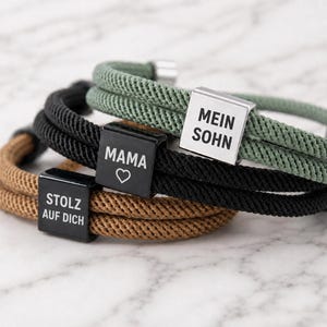 Può includere: Tre bracciali a strati realizzati in corda intrecciata color marrone chiaro, nero e verde salvia. Ogni bracciale presenta una perlina quadrata in metallo con testo inciso. La perlina marrone riporta STOLZ AUF DICH, quella nera MAMA con un cuore.