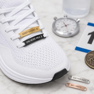 Könnte beinhalten: Ein weißer Sportschuh mit zwei Metall-Schnürsenkelplaketten, eine goldene mit dem Text KEEP GOING und einem Sternsymbol, eine schwarze mit dem Text ONE MORE MILE und einem Läufersymbol. Zwei weitere silberne und roségoldene Plaketten zeigen RUN YOUR RACE und NO LIMITS.