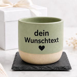 Può includere: Una tazza in ceramica color verde salvia e beige con il testo nero stampato "dein Wunschtext" e una piccola icona a forma di cuore nero centrata sul davanti. La tazza presenta una finitura opaca liscia e una forma cilindrica con base piatta.