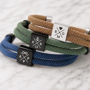 Könnte beinhalten: Drei geflochtene Kordelarmbänder in Blau, Grün und Beige. Jedes verfügt über einen quadratischen Metallschieber mit einer Kreuzgravur. Das blaue Armband zeigt ein Unendlichkeitssymbol, das grüne einen Anker und 23, das beige ein Herz. Die Buchstaben A, P, K, R, B, D und M sind ebenfalls eingraviert.