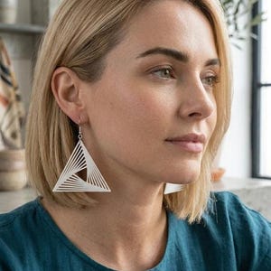 Peut inclure: Boucles d'oreilles triangulaires géométriques blanches avec un design en couches. Les boucles d'oreilles sont suspendues à des crochets de couleur argentée. La femme porte un haut de couleur sarcelle.