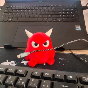 Peut inclure: Une petite figurine de monstre rouge vif avec des cornes, de grands yeux blancs et une expression renfrognée, tenant un câble de charge tressé noir et blanc dans sa bouche, posée sur un tapis de bureau noir devant un clavier d'ordinateur.