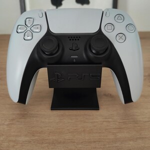 Peut inclure: Une manette blanche et noire PlayStation 5 sur un support noir. Le support affiche "PS5". La manette a un design symétrique avec des joysticks et des boutons noirs, typique des manettes modernes.